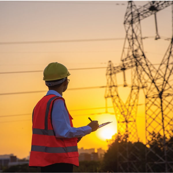 Utility worker in safety gear inspecting power lines at sunset with a clipboard.