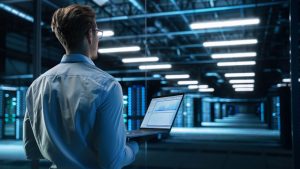 A person in a data center, holding a laptop and wearing glasses, gazes at server racks.