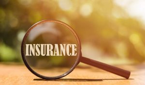 Magnifying glass focusing on the word INSURANCE on a wooden surface with a blurred sunny background.